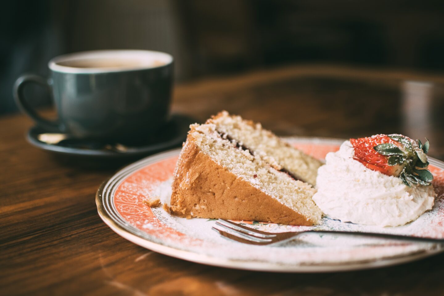 A black cup and saucer with hot drink on a wooden table beside a pink and white patterned plate upon which is a slice of cake with cream and a piece of strawberry 