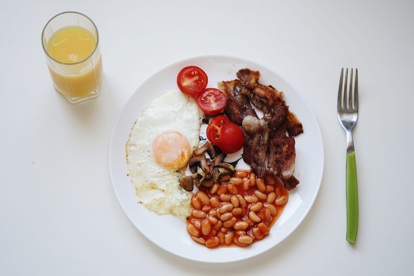 White table with an aerial view of a half glass of orange juice to the top left of a round white plate containing 4 baby tomato halves, a fried egg, some beans, cooked mushrooms, and several slices of bacon, with a green handled silver fork to the right of the plate