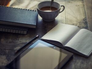 A half full coffee cup with spoon on a wooden floor with several ring bound notebooks, one open one and an electronic notebook and pencil in front of it.