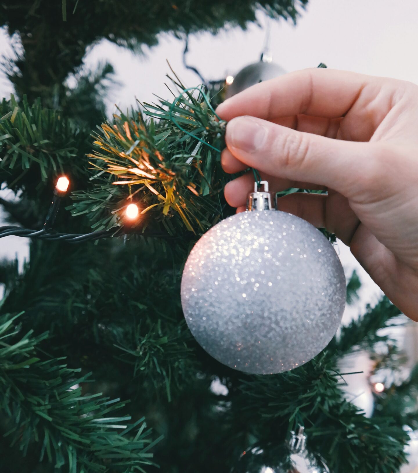 Partial picture of a green Christmas tree with a hand hanging up a silver bauble